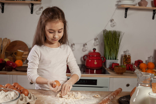 Gyönyörű kislány öltözött fehér blúz van gyártás egy tésztát kenyérsütés a konyhában. - Fotó, kép