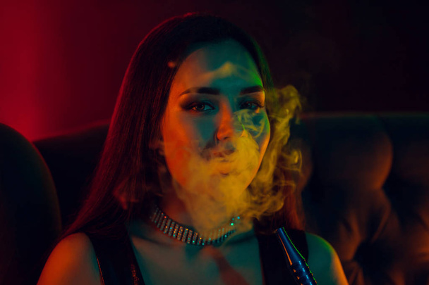 Сексуальная модель брюнетки курит кальян, выдыхая сигарету в роскошном ночном клубе
. - Фото, изображение