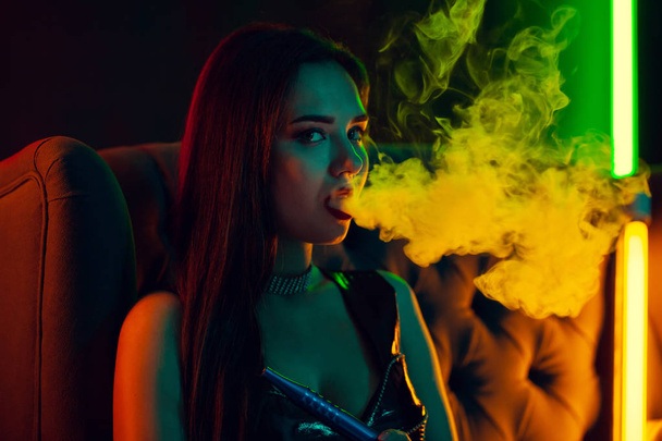 Sexy brunette model is smoking a hookah exhaling a smoke at a luxury night club. - Фото, зображення