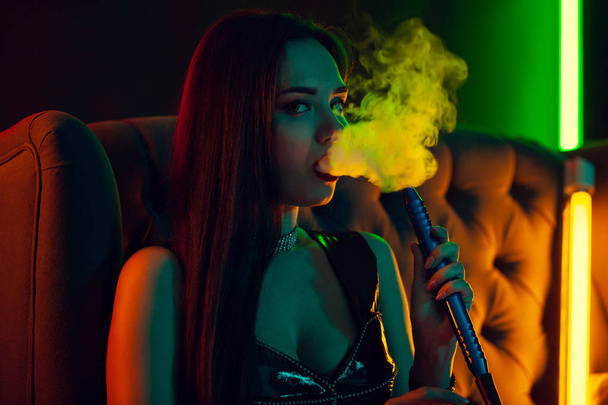 Sexy brunette model is smoking a hookah exhaling a smoke at a luxury night club. - Foto, Imagem