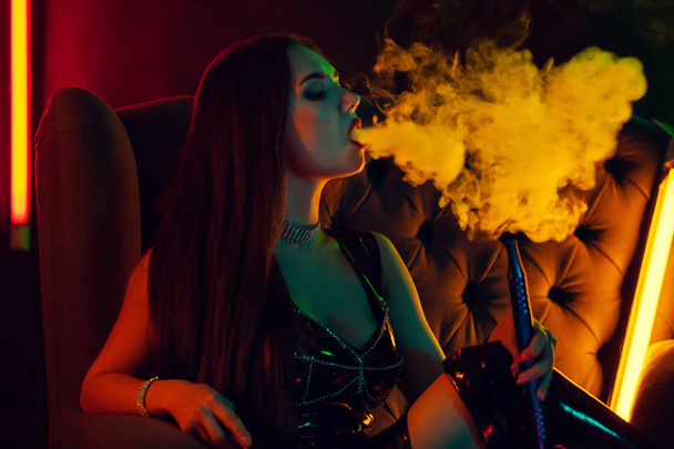 Seksowny brunetka model jest palenie a Hookah wydechu a smoke w a luksusowy klub nocny. - Zdjęcie, obraz