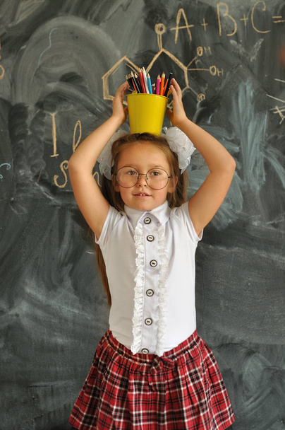 Portrait of a happy school girl standing with yellow bucket with colored pencils on head.Go to school for the first time. - Photo, Image
