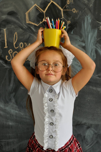 Portret szczęśliwej uczennice stojący z żółtym wiadrem z kolorowymi ołówki na głowie. Idź do szkoły po raz pierwszy. - Zdjęcie, obraz