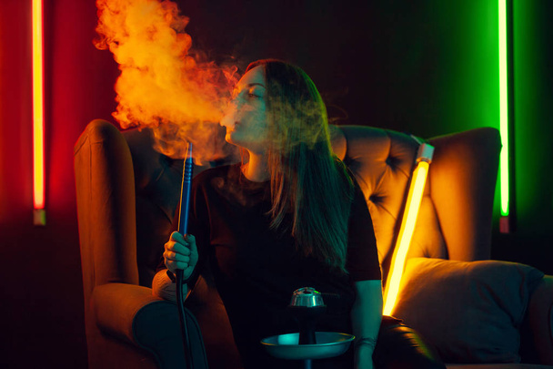 Pretty girl in a black t-shirt is smoking a hookah exhaling a smoke at a luxury night club. - Фото, зображення