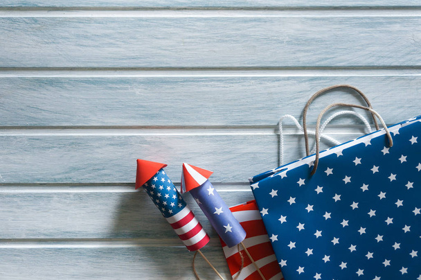 4th July holiday shopping concept. Shopping bags in national american colors and firework rockets over wooden background - 写真・画像