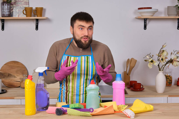 Młody, przystojny brodaty mężczyzna w kuchni, pokazuje całą swoją ekipę sprzątającą - detergenty, szczotki, spraye. Myśli, że jest gotowy na prawdziwe sprzątanie. - Zdjęcie, obraz