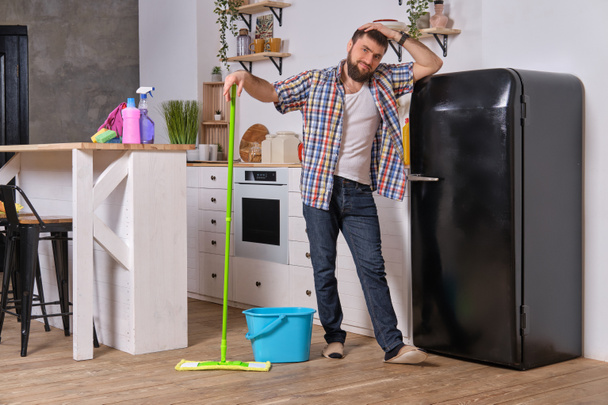 El ama de llaves del marido limpia la cocina. Joven hombre feliz con fregona verde lava el suelo
 - Foto, imagen