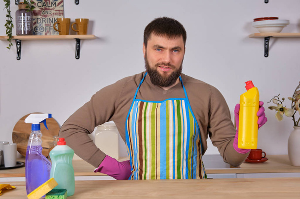 Young bearded man in kitchen makes advertising. He promotes detergent, holding it in one hand and shows at camera - 写真・画像