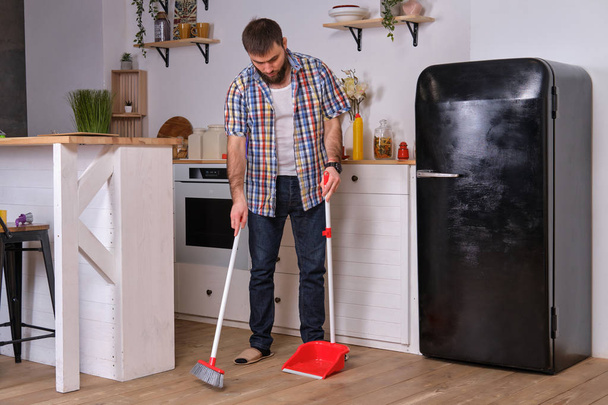 Young handsome bearded man in a kitchen, wearing a plaid shirt, sweeping the floor with a shovel and broom. - 写真・画像