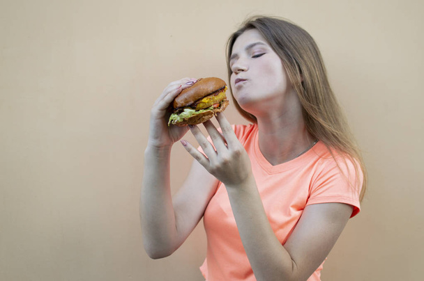 çekici genç kız turuncu bir t-shirt turuncu duvara karşı duruyor ve elinde büyük bir burger tutar, o ağzını geniş açar ve yiyor - Fotoğraf, Görsel