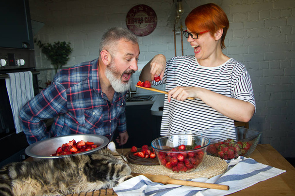 erwachsenes Paar Mann und Frau schälen und schneiden Erdbeeren für Erdbeermarmelade, füttern einander, lachen und haben Spaß, das Maine Coon Kätzchen steht auf dem Küchentisch - Foto, Bild