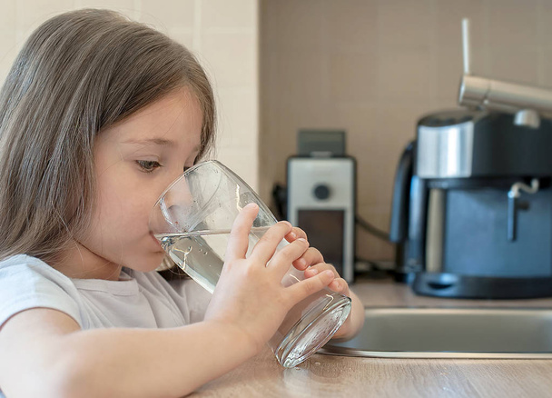 Little child is drinking clean water at home, close up. Caucasian cute girl with long hair is holding a water glass in her hands. Taking care of own health. Concept of healthy lifestyle, good habit - Foto, imagen