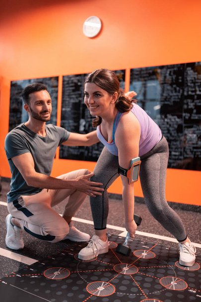 Женщина с ожирением улыбается тренируясь в тренажерном зале с тренером
 - Фото, изображение