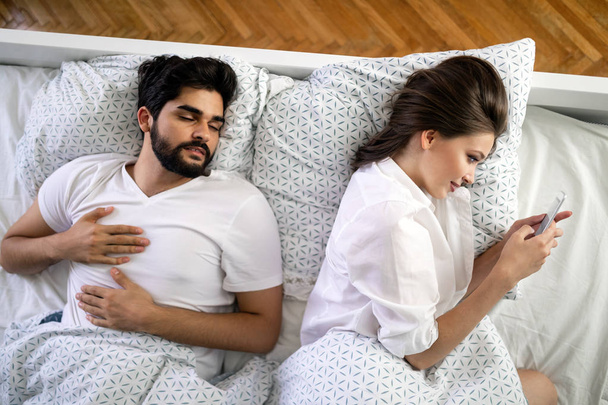 Beautiful young wife texting with lover on smartphone while husband sleeping - Foto, Imagem