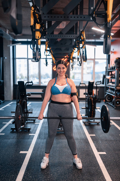 Donna grassoccia che tiene il bilanciere in piedi nel centro fitness
 - Foto, immagini
