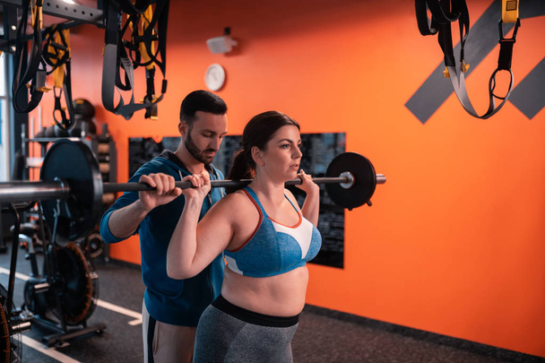 Bearded dark-haired trainer helping plump woman to lift barbell - Photo, Image