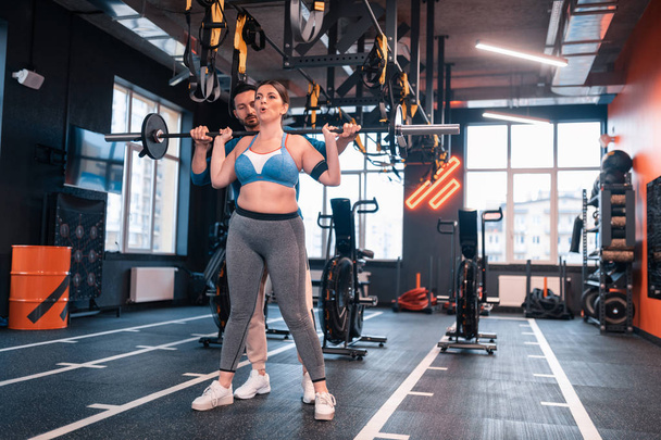 Woman hardly breathing after lifting barbell standing near trainer - Foto, afbeelding