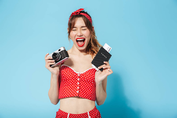 Young pin up pretty summer girl in swimwear posing isolated over blue background holding camera and passport with tickets. - 写真・画像