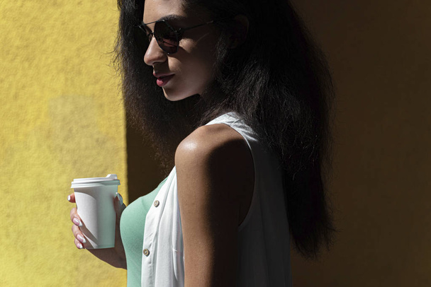 Attractive young woman with paper coffee cup standing against yellow wall - Fotó, kép