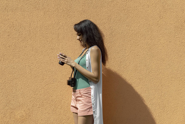Bájos fiatal nő retro kamerával állva fényében narancssárga fal - Fotó, kép