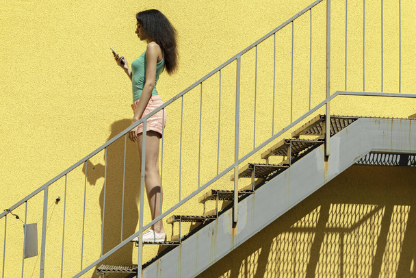 Beautiful young woman with cellphone standing on stairs against yellow wall - Фото, зображення