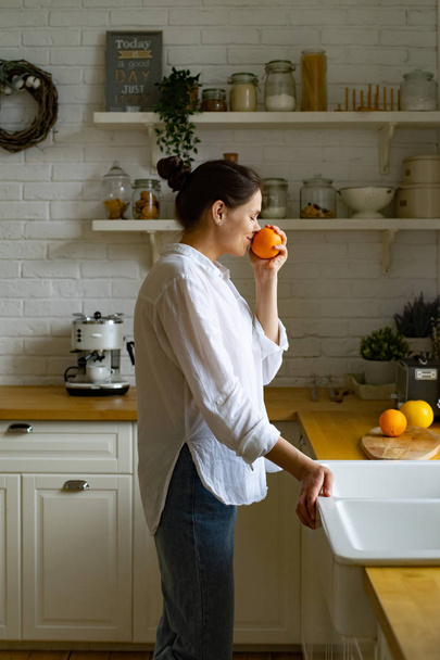 Jeune femme sentant l'orange tout en restant debout à la cuisine
  - Photo, image