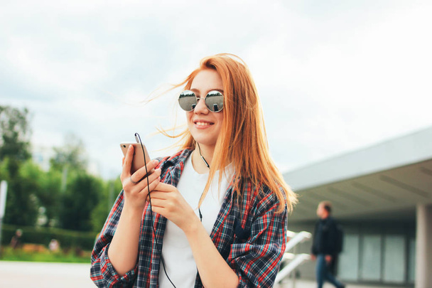 Atractiva pelirroja sonriente en gafas de sol redondas con teléfono en sus manos en ropa casual escuchando música en la calle de la ciudad
 - Foto, Imagen