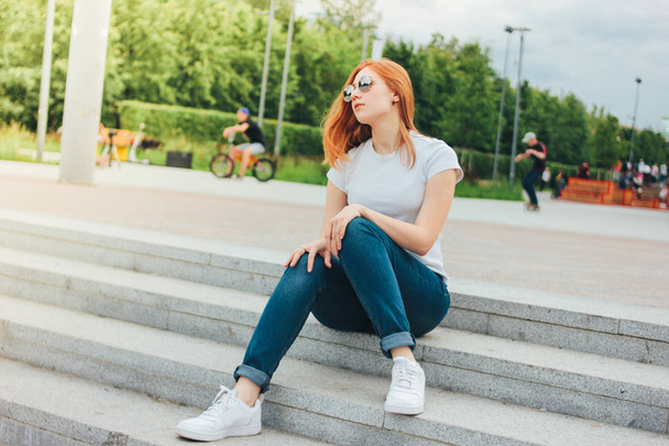 Attractive redhead smiling girl in round sunglasses in casual clothes sitting on street in city - Fotó, kép