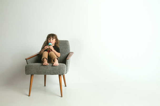 Criança sentada e brincando em poltrona no fundo do estúdio branco
 - Foto, Imagem