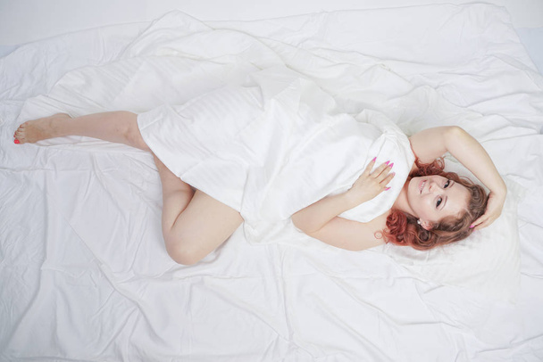 kaunis nuori tyttö makaa valkoisella sängyllä ja nauttii aamusta. viehättävä valkoihoinen nainen on onnellinen ja rentouttava. unelias ja laiska henkilö hitaasti poseeraa yksin
. - Valokuva, kuva