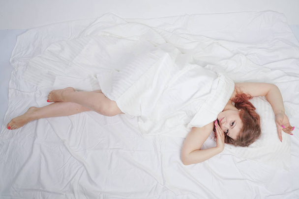schöne junge Mädchen liegt auf einem weißen Bett und genießt den Morgen. Die charmante Kaukasierin ist glücklich und entspannt. schläfriger und fauler Mensch posiert langsam allein. - Foto, Bild