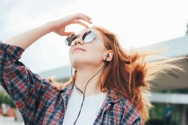 Привлекательная рыжая улыбающаяся девушка в круглых солнцезащитных очках с телефоном в руках в повседневной одежде слушает музыку на улице в городе
 - Фото, изображение