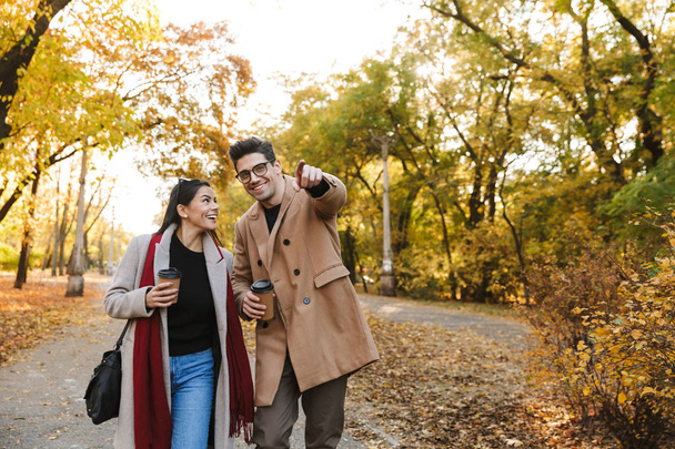 Portrait of happy couple drinking takeaway coffee from paper cups while walking in autumn park - 写真・画像