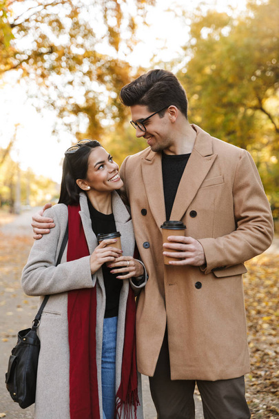Portrait of young couple drinking takeaway coffee and looking at each other while walking in autumn park - Foto, imagen