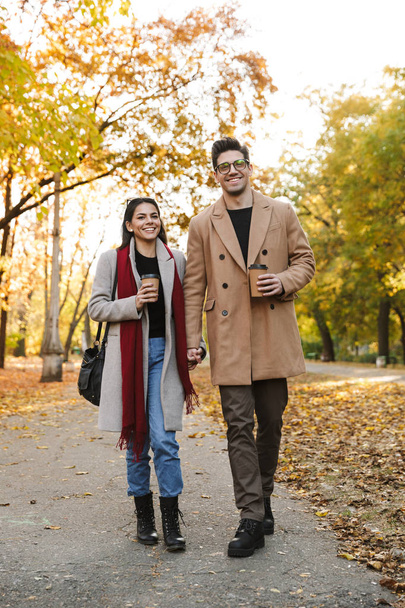 Portrait of casual couple drinking takeaway coffee from paper cups while walking in autumn park - Фото, зображення