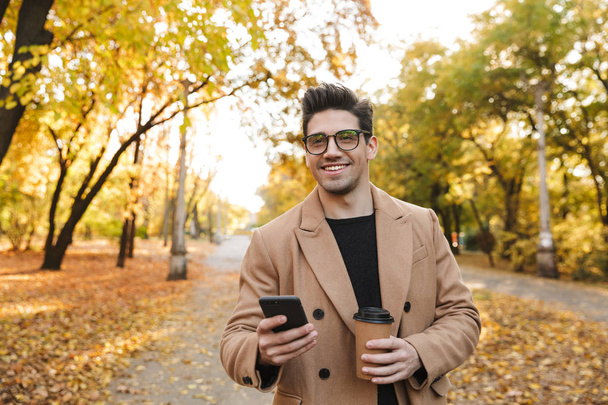 Image of handsome european man using cellphone and smiling in autumn park - Foto, imagen