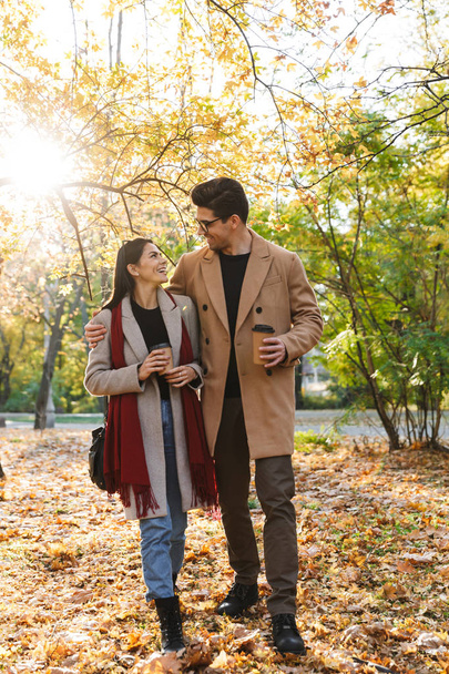 Portrait of beautiful couple drinking takeaway coffee and looking at each other while walking in autumn park - Foto, immagini