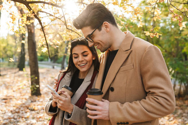 Portrait of stylish couple drinking takeaway coffee and looking at smartphone while walking in autumn park - Фото, зображення