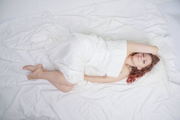 όμορφο νεαρό κορίτσι είναι ξαπλωμένο σε ένα λευκό κρεβάτι και απολαμβάνοντας το πρωί. γοητευτική καυκάσια γυναίκα είναι ευτυχισμένη και χαλαρωτική. νυσταγμένη και τεμπέλη που ποζάρει αργά μόνη. - Φωτογραφία, εικόνα