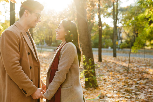 Imagen de pareja joven y alegre sonriendo y tomándose de la mano en el parque de otoño
 - Foto, imagen