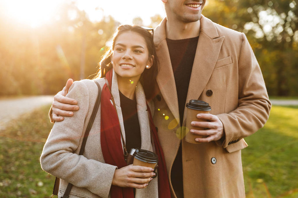 Portrait of attractive couple drinking takeaway coffee from paper cups while walking in autumn park - Foto, imagen