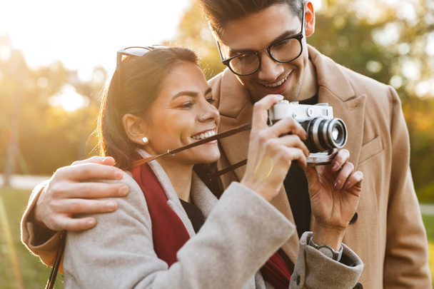 Portrait of smiling couple taking photo on retro camera while walking in autumn park - Photo, Image