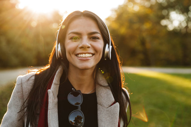 Portrait of young brunette woman wearing coat listening to music with headphones and smiling in autumn park - Photo, Image