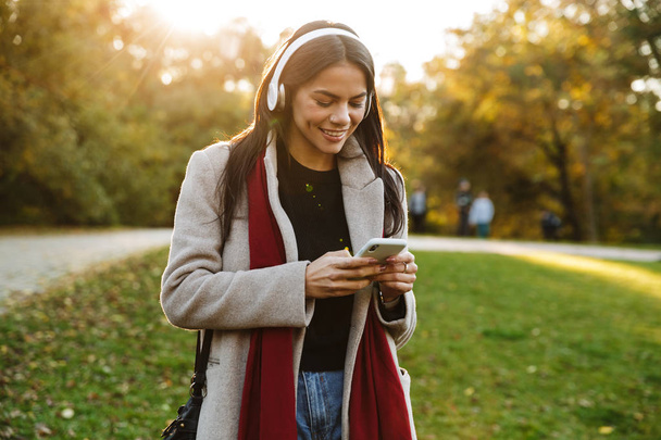 Portrait of happy nice woman wearing coat listening to music with headphones and using cellphone in park - Photo, image