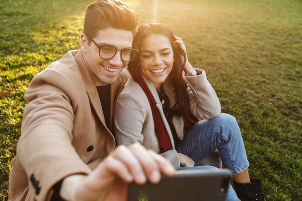 Image of cute european couple taking selfie on cellphone while sitting on grass in park - Foto, immagini