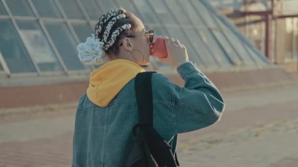 Camera follows young woman with backpack walking at sunset outdoors. Attractive young female with sunglasses drinking coffee or tea outdoors. Medium shot - Filmmaterial, Video