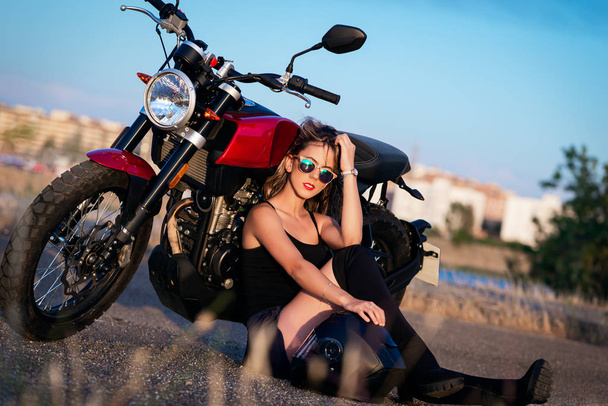 Молодая красивая женщина, сидящая на земле рядом с Nake Racer motorbi
 - Фото, изображение