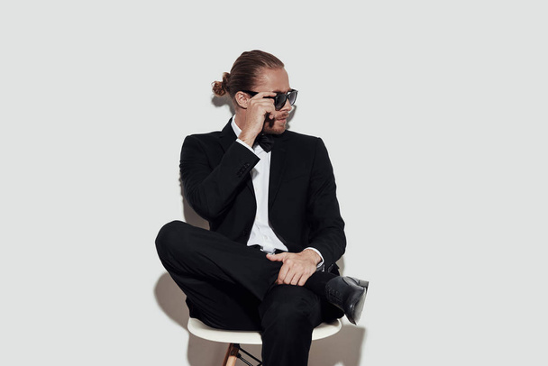 Confident and ahndsome. Charming young man in full suit looking away and adjusting sunglasses while sitting against grey background - Photo, Image