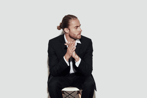 Moment to overthink everything. Handsome young man in full suit looking away and keeping hands clasped while sitting against grey background - Photo, Image