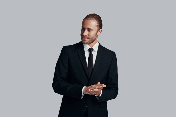 Ready to do business. Handsome young man in full suit looking away and keeping hands clasped while standing against grey background - Photo, Image